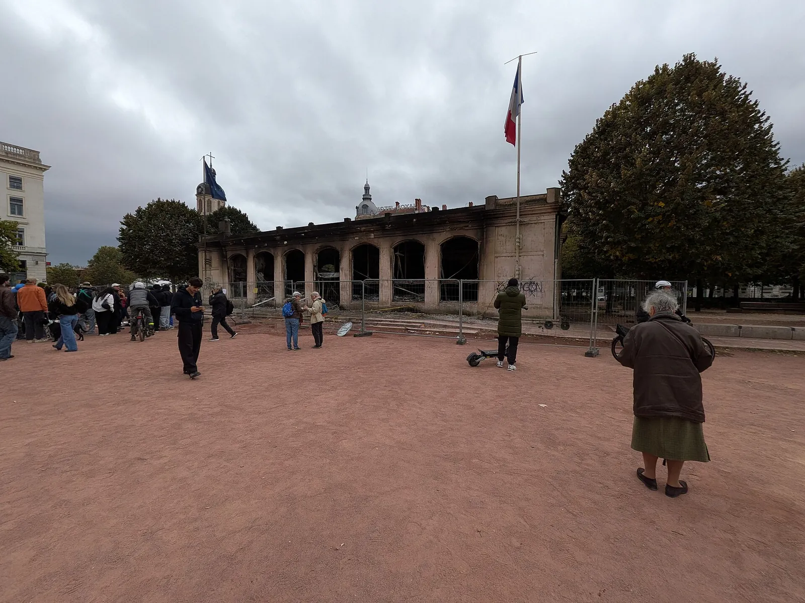 Place Bellecour à Lyon le lendemain du 17 octobre 2026 : le pavillon de l'office de tourisme noirci, entouré de barrières, avec des passants